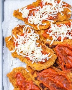 Breaded chicken cutlets on a baking sheet, topped with marinara sauce and shredded mozzarella, ready for the oven.