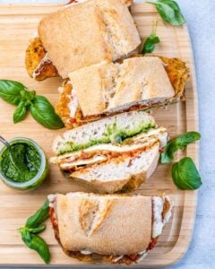 Sliced Chicken Parmesan Sandwiches served on a wooden board with pesto sauce and fresh basil scattered around.