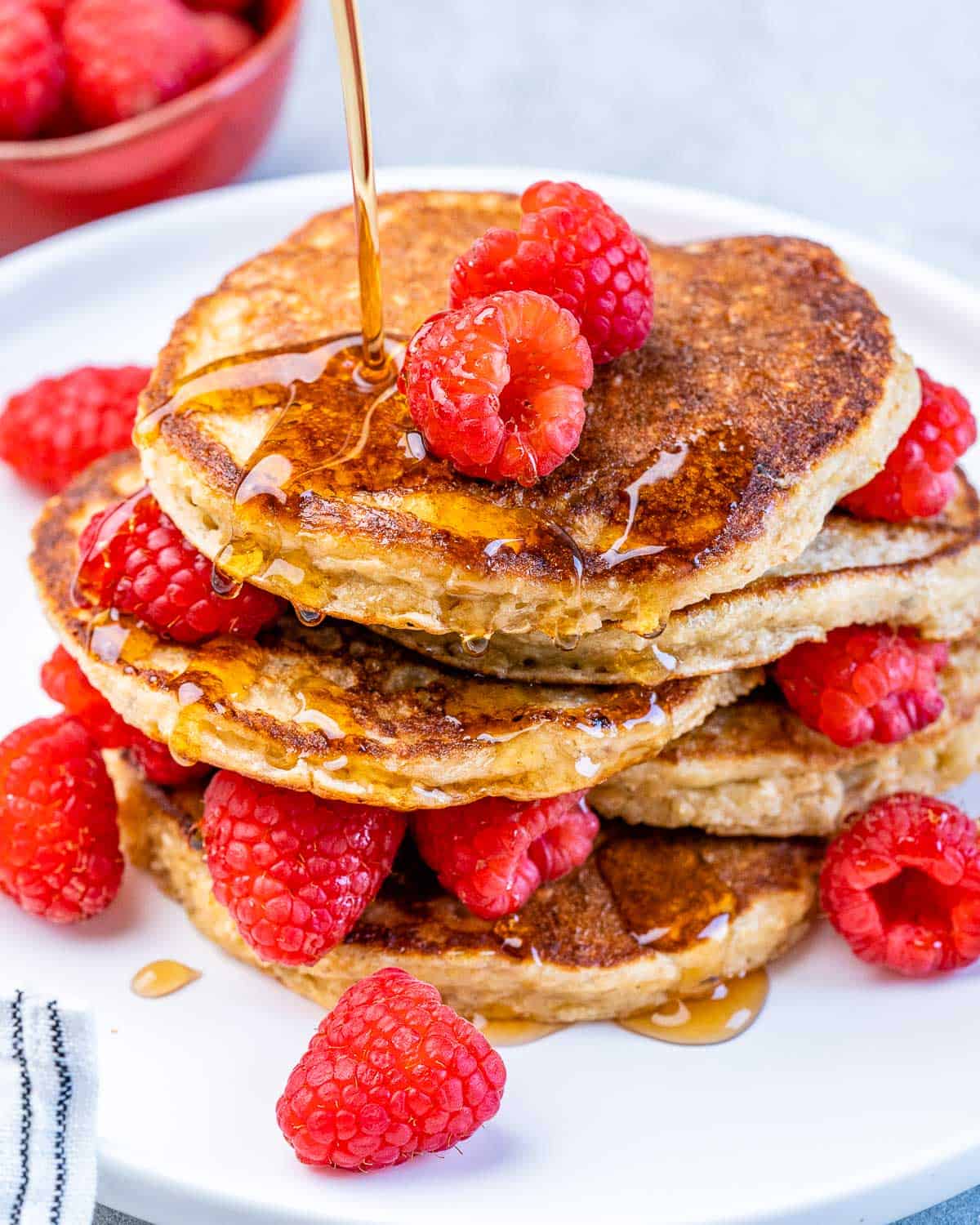 Drizzling syrup over pancakes that are topped with raspberries.