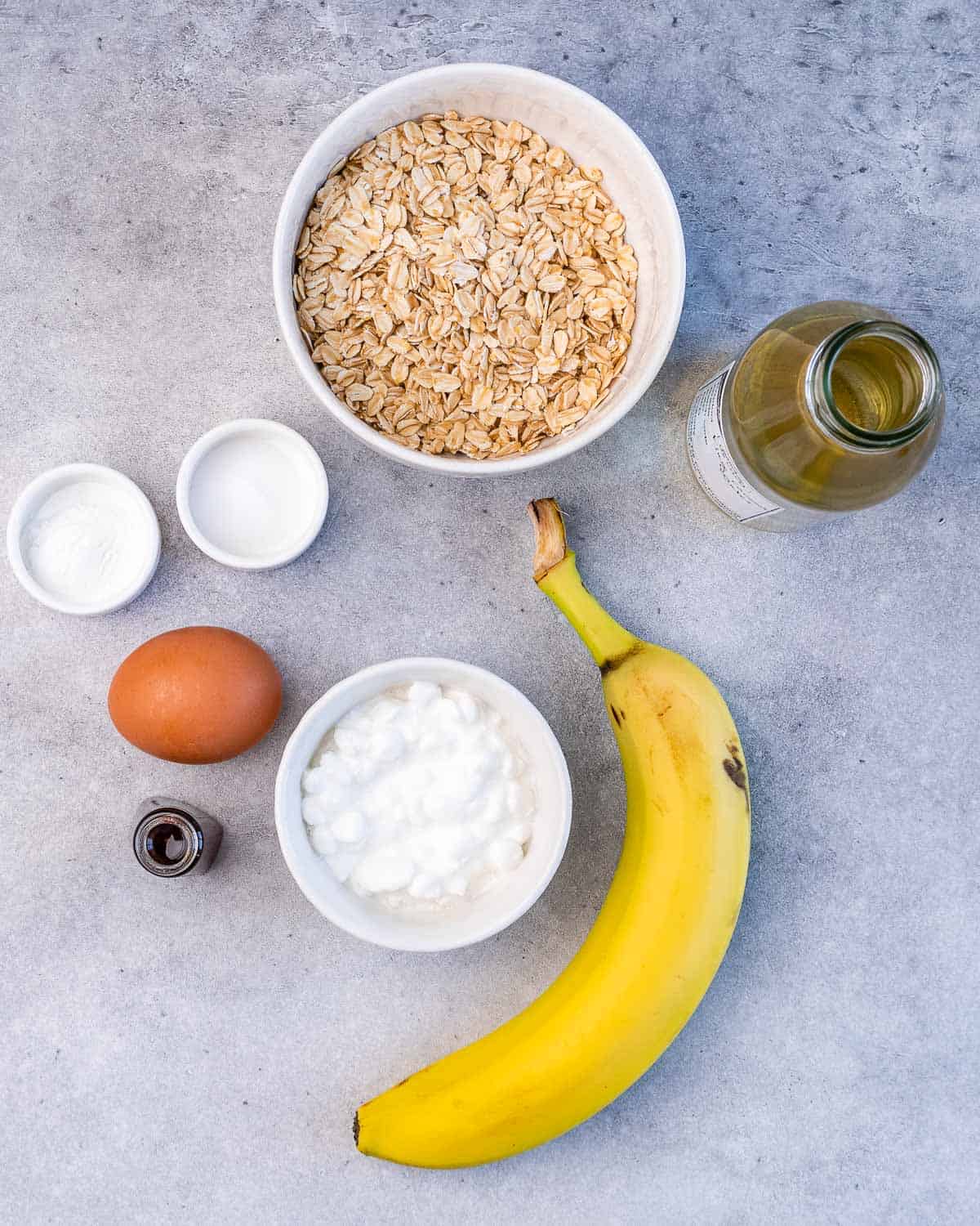 Bowl of rolled oats near an egg, banana, bowl of cottage cheese and vanilla. 