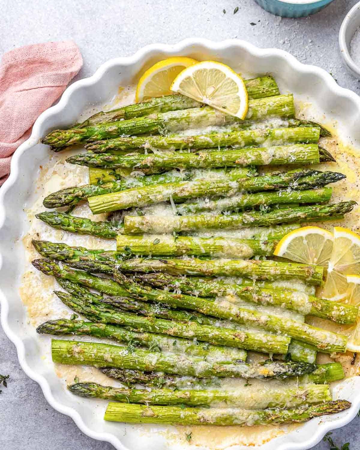baked asparagus in a dish topped with parmesan cheese and lemon garnishes.