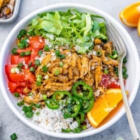 A round bowl with greens, rice, and tomatoes, topped with shredded chicken carnitas.