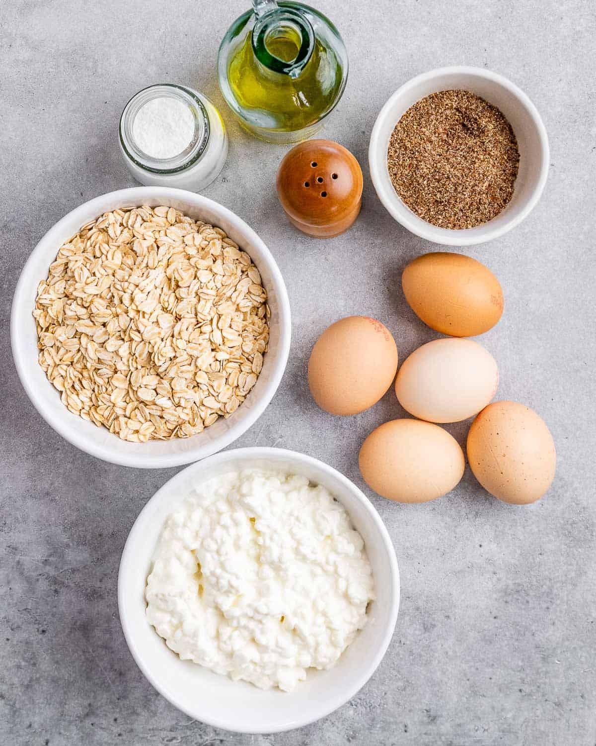 ingredients to make cottage cheese bread laid out. They are: cottage cheese in a bowl, rolled oats in a bowl, olive oil, baking powder, salt, ground flax seed in a bowl, and 6 eggs.