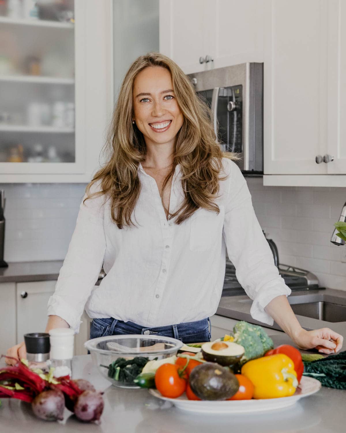 Ella Davar in a white shirt standing by the counter in a kitchen with fruits on the counter.