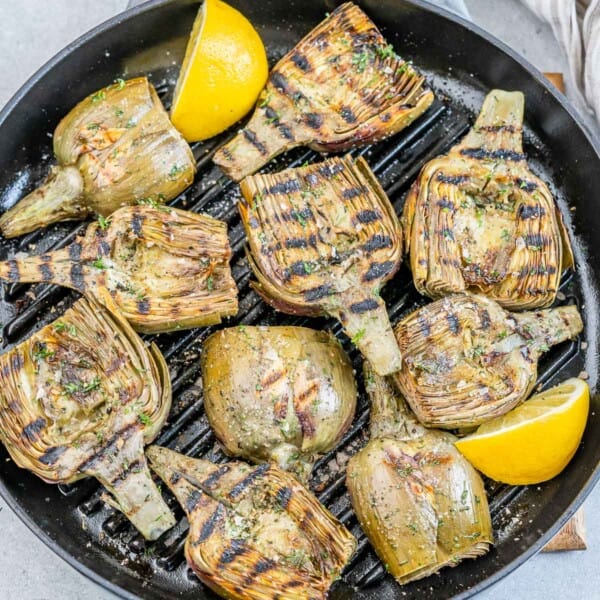 A grill pan with grilled artichoke halves with lemon garnishes.