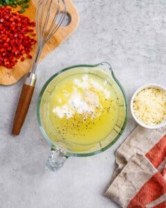 Overhead shot of a glass mixing bowl with egg whites, cottage cheese, and seasoning, surrounded by chopped red pepper and shredded cheese on a light surface.