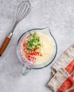 Mixing bowl with egg white mixture, red pepper, green onion, and shredded cheese, with a metal whisk resting inside.