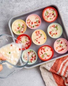 Muffin tin filled with unbaked egg white mixture, visible bits of red pepper and green onion floating on top.