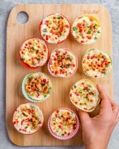 Hand placing a silicone muffin cup filled with egg white mixture onto a wooden board with other baked egg white bites.