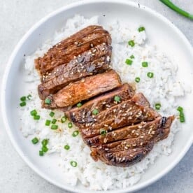 A round white plate with white rice topped with sliced teriyaki steak garnished with chopped green onions and sesame seeds.