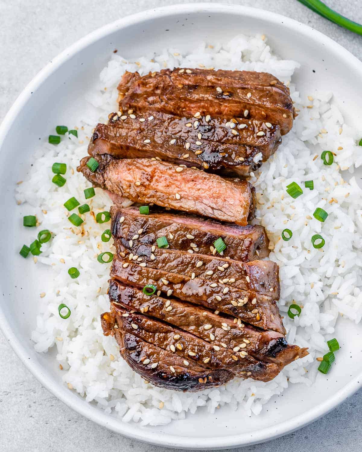 A round white plate with white rice topped with sliced teriyaki steak garnished with chopped green onions and sesame seeds.