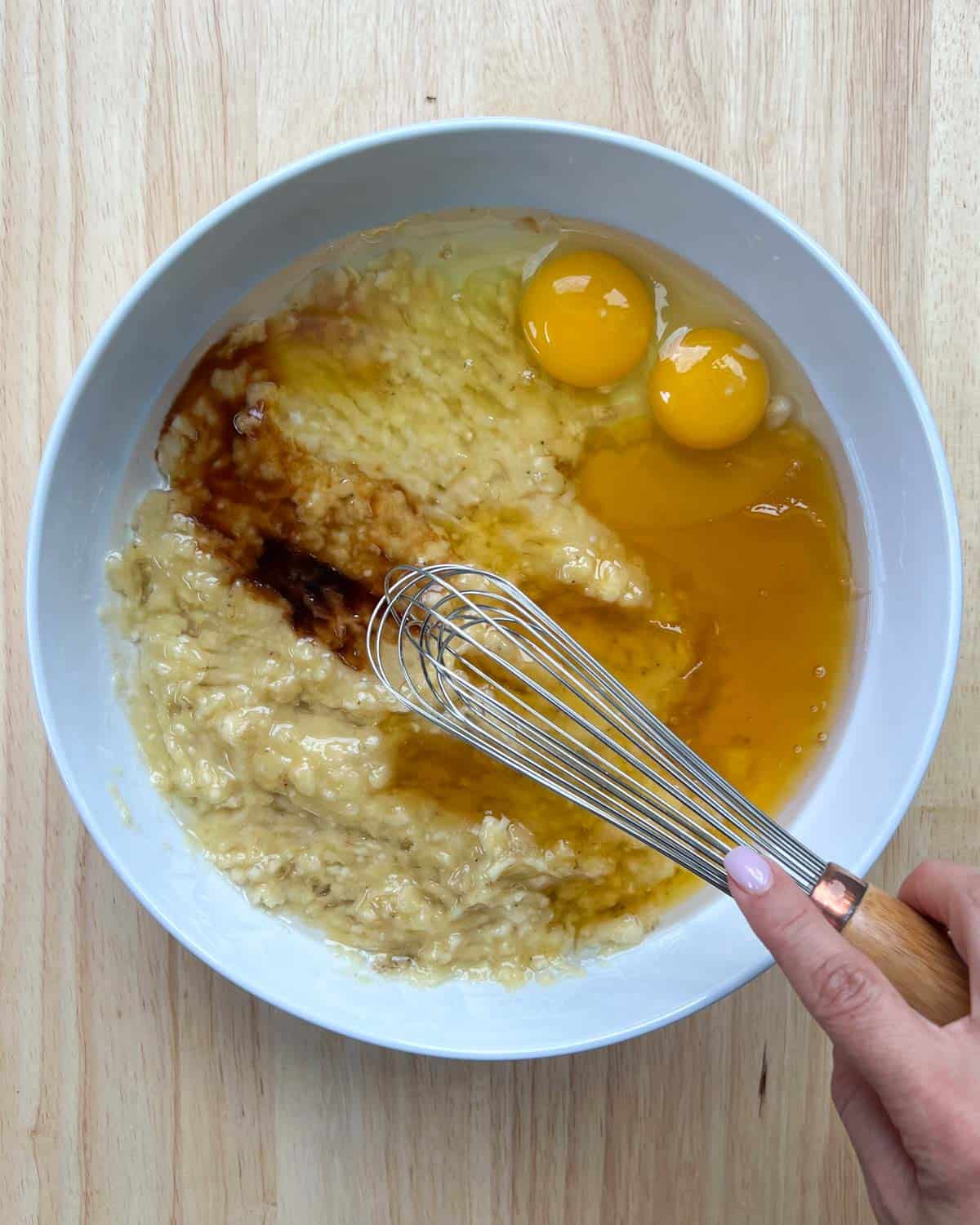 eggs, mashed bananas, vanilla, honey, and coconut oil in a white bowl with hand holding whisk over it.