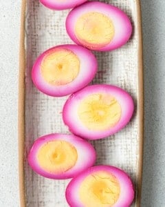Multiple halved pink pickled eggs laid out in a row on a rectangular tray lined with textured cloth or parchment.