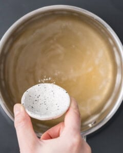 Large stainless steel pot containing warm golden-brown pickling brine with a hand adding in ingredients from a shallow dish.