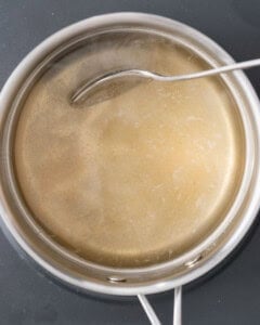 Large stainless steel pot containing warm golden-brown pickling brine being stirred with a white spoon.