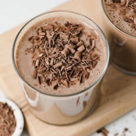 A clear cup on a cutting board filled with coffee smoothie and topped with shaved chocolate.