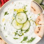 a round bowl of tzatziki sauce topped with sliced cucumber and a few pita bread slices in dip.