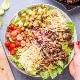 A hand holding a bowl of salad that's burger style with minced meat, lettuce, tomatoes, and pickles.