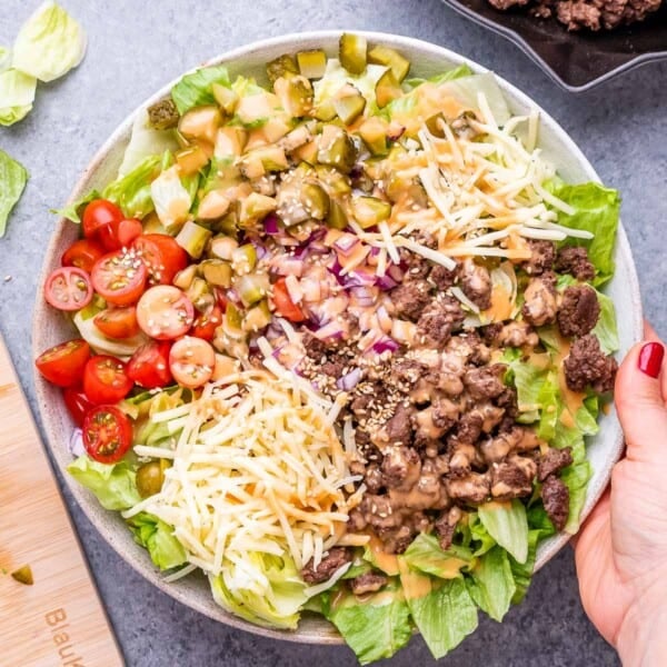 A hand holding a bowl of salad that's burger style with minced meat, lettuce, tomatoes, and pickles.