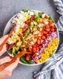 A hand serving up a portion of the Cobb salad with wooden tongs, showing the layers of toppings being tossed together.