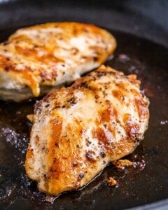 Two golden, seared chicken breasts cooking in a skillet, seasoned and cooked until juicy and browned on the outside.