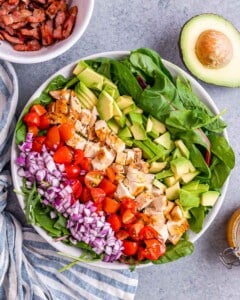 Top-down view of the salad before dressing, with bold stripes of avocado, tomato, red onion, chicken, bacon, feta, and corn on a bed of spinach.