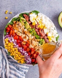 Dressing being poured onto the fully assembled Cobb salad in a large bowl, with every topping clearly visible.