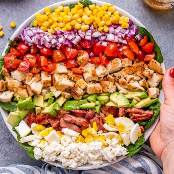Top view of a colorful Chick Fil A Cobb Salad with grilled chicken, corn, tomatoes, avocado, red onion, feta, and turkey bacon, all arranged in neat rows on a bed of greens with a hand holding the bowl.