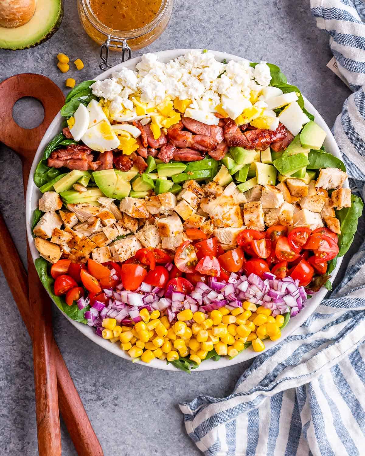 Close-up of a full Cobb salad arranged in stripes of vibrant toppings over spinach, with wooden salad tongs on the side.