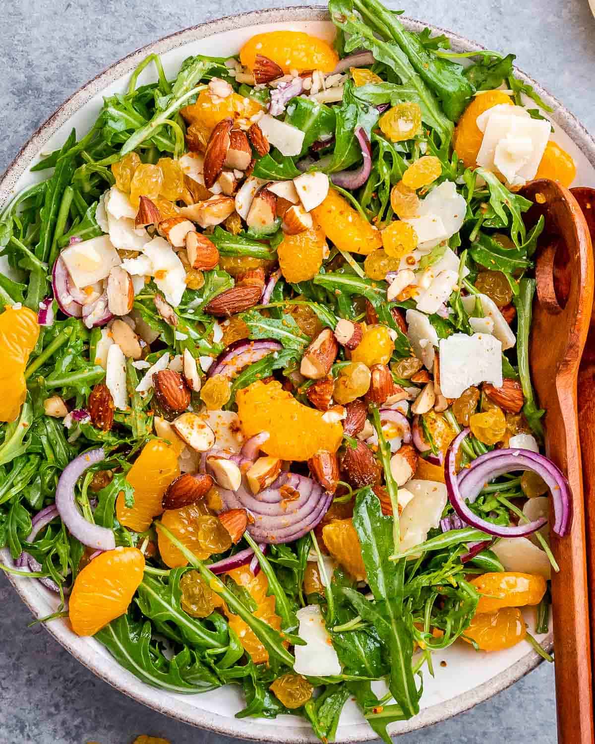 Large white bowl filled with arugula, topped with mandarin segments, sliced red onion, raisins, almonds, and parmesan cheese.
