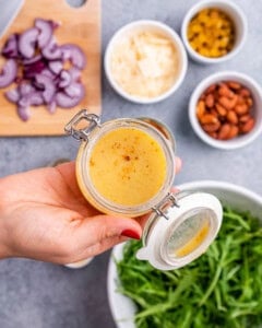 Close-up of the homemade dressing in a small jar with the lid open, held above a counter with salad ingredients in the background.