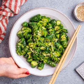 Hand holding a round bowl with cucumber salad topped with sesame.