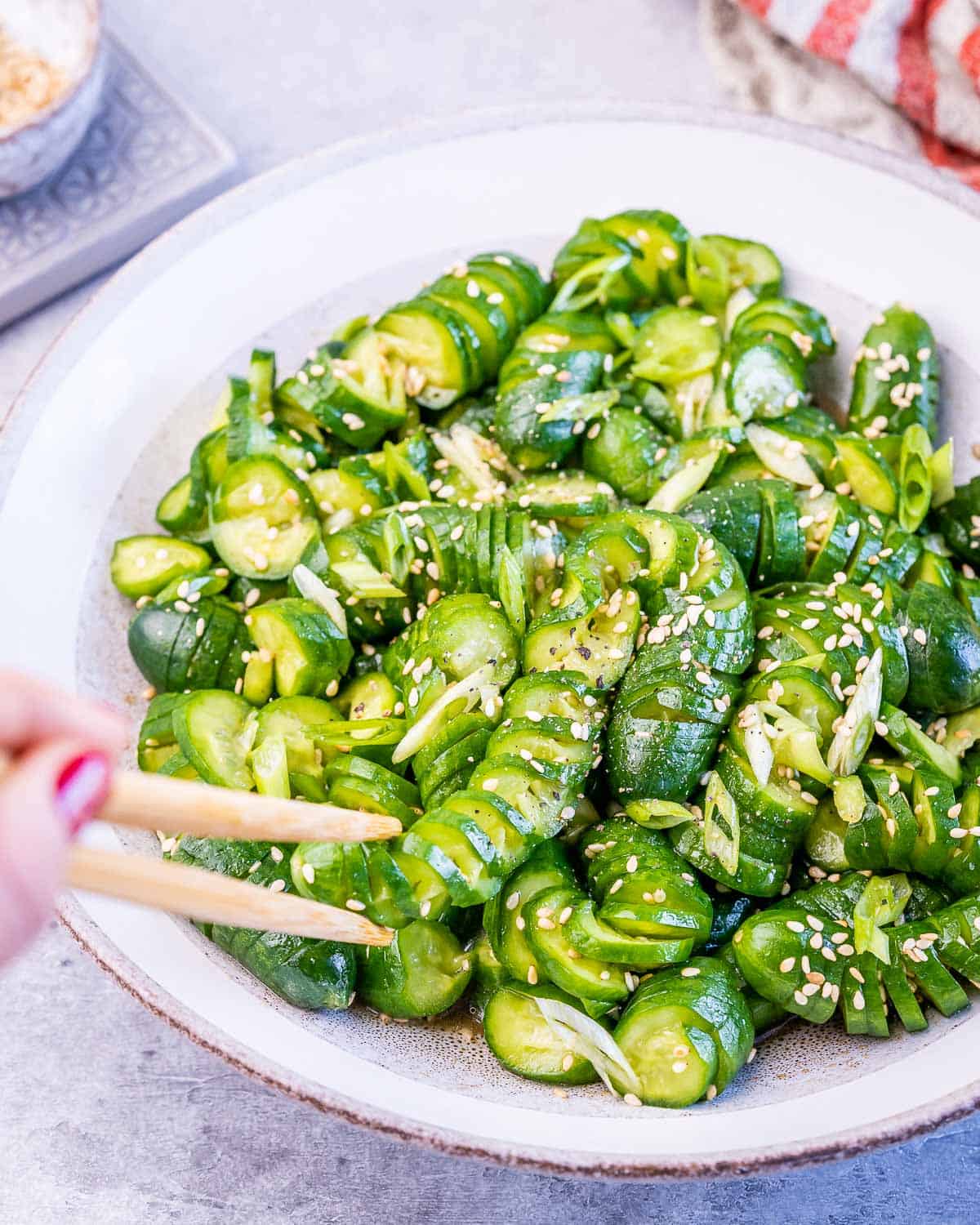 chopsticks grabbing cucumber salad on a white plate topped with sesame seeds.