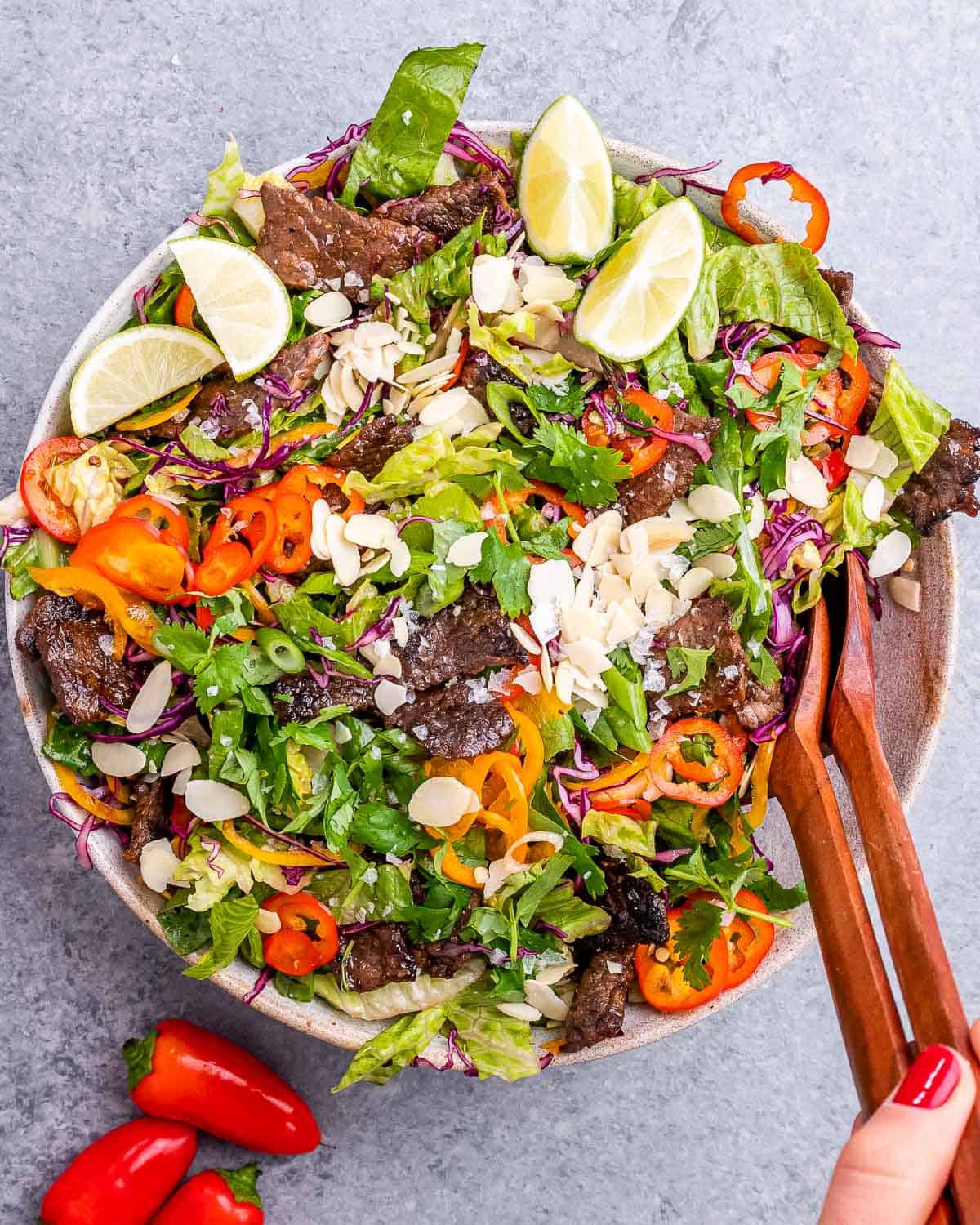 A white bowl of Thai beef salad with wooden tongs.