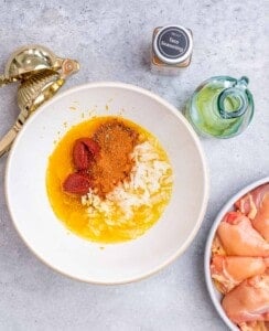 Overhead view of a mixing bowl filled with marinade ingredients including orange juice, garlic, and spices, next to raw chicken.