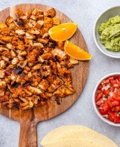Chopped grilled chicken on a wooden board with orange wedges, guacamole, and pico de gallo nearby.