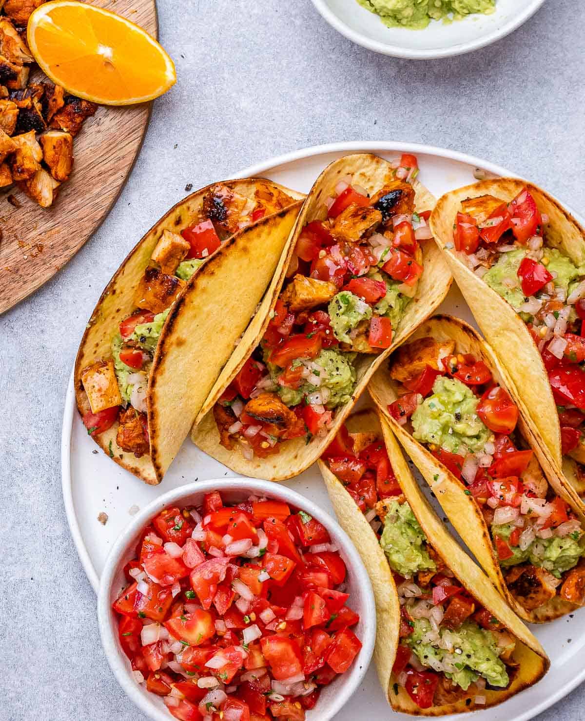 Plate of three tacos surrounded by bowls of diced tomatoes and grilled chicken, ready to serve.