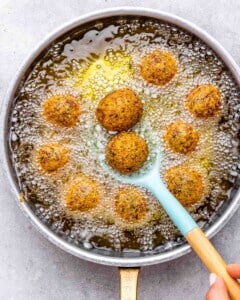 Falafel balls frying in hot oil inside a deep skillet with a slotted spoon lifting one out.