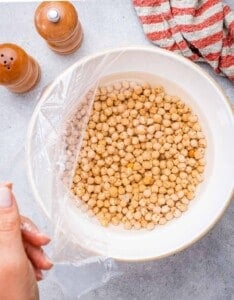 Bowl of dried chickpeas soaking in water, covered with plastic wrap.