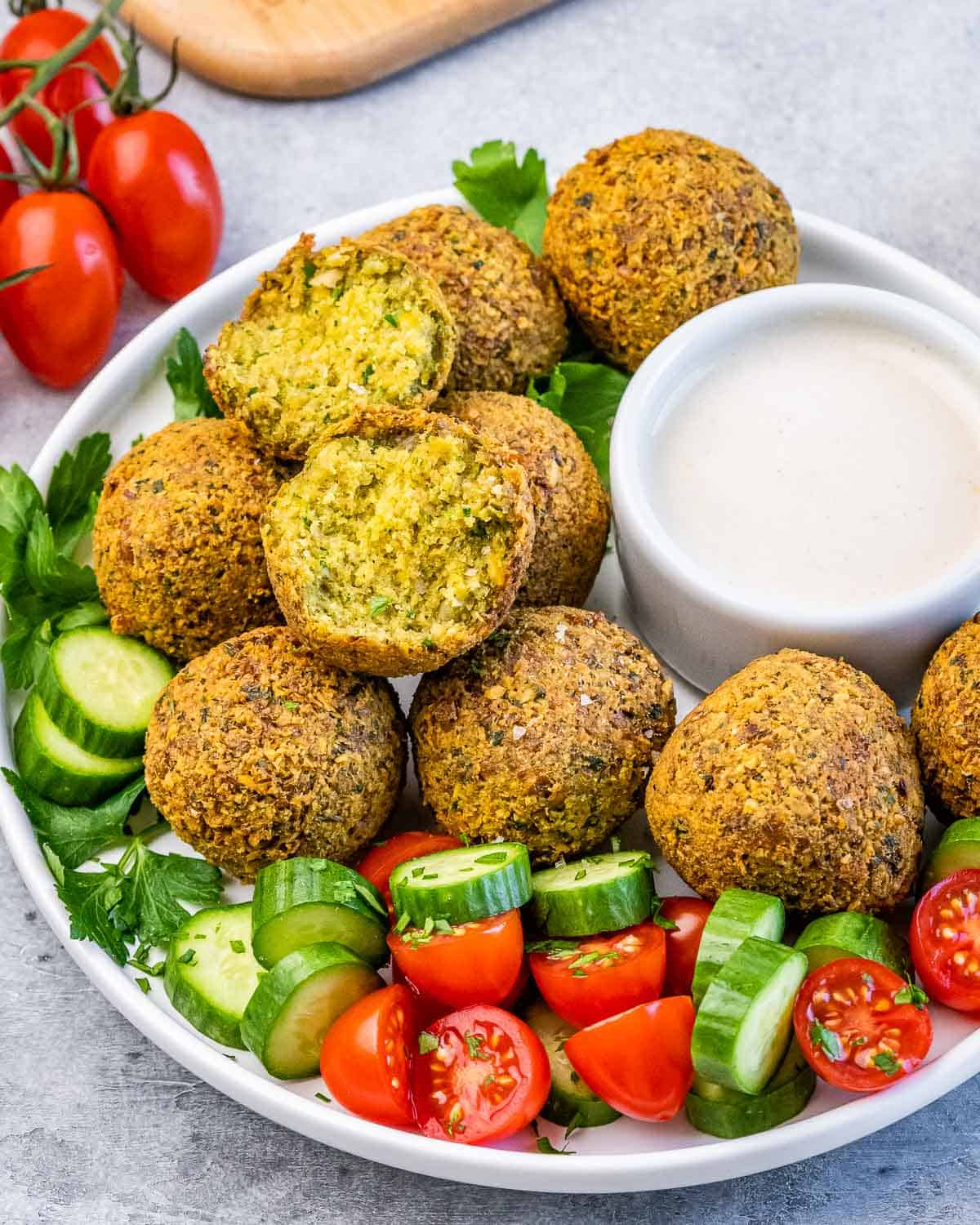 Falafel balls served with cucumber, tomato, parsley, and a creamy dipping sauce on a round white plate.