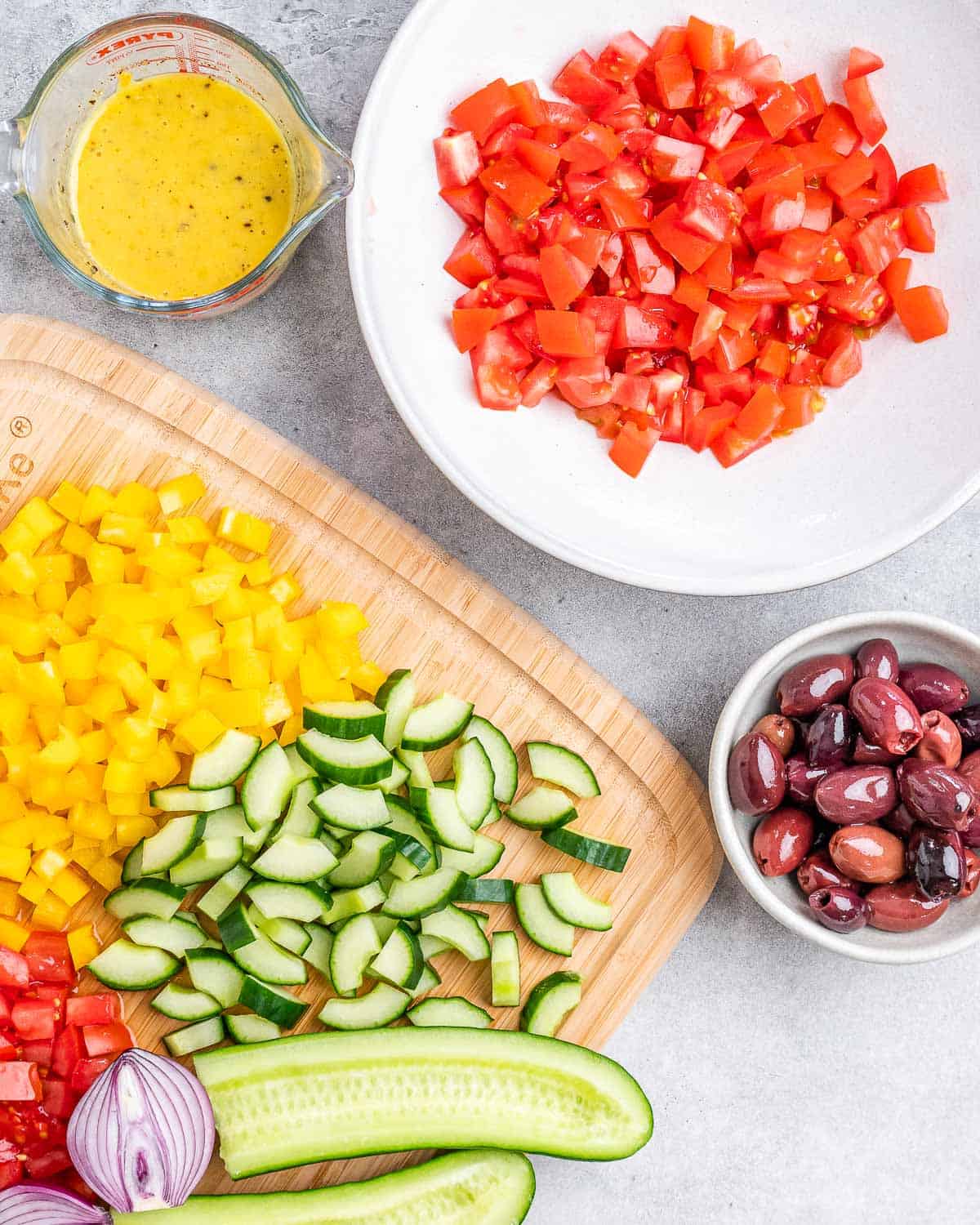 Chopped tomatoes in a bowl alongside dressing, chopped cucumber, olives and chopped yellow bell pepper.