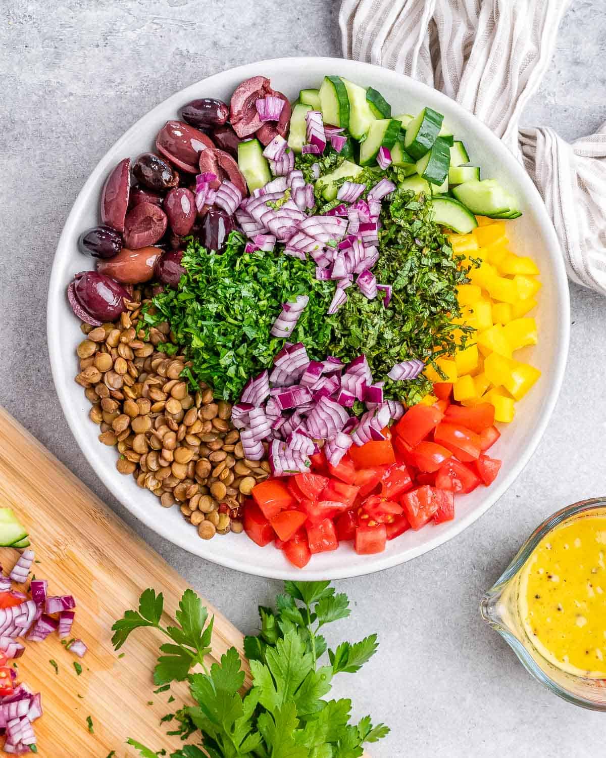 Lentil salad ingredients added to a white bowl near a glass filled with yellow dressing.