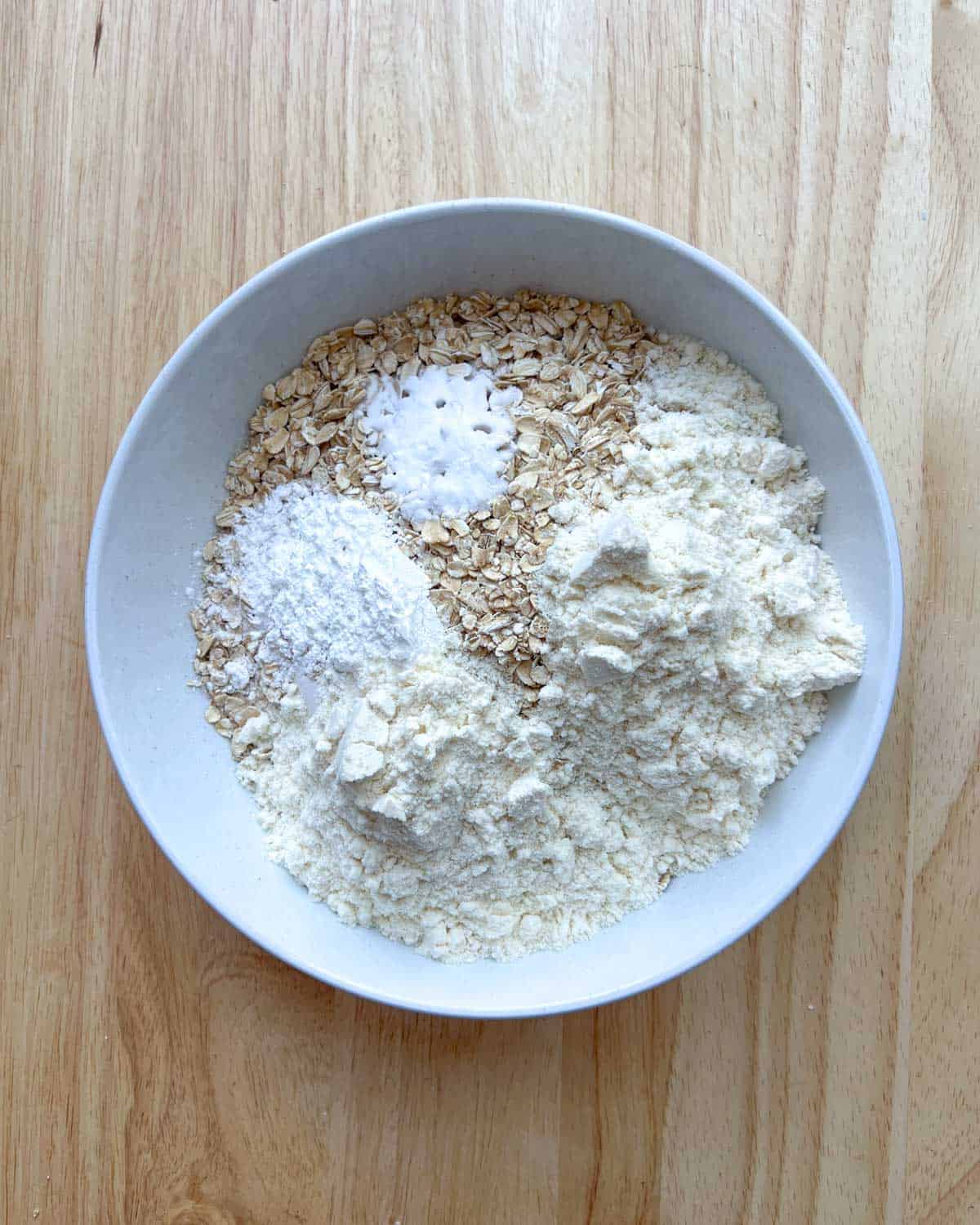 oats, flour, baking powder, baking soda, salt, and protein powder, added to a bowl.