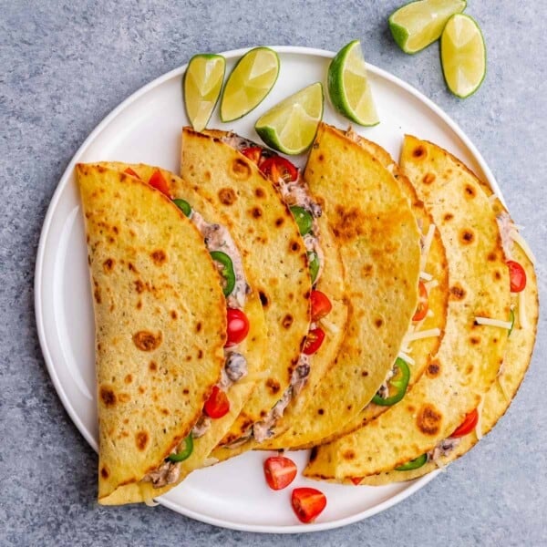 A close-up of four folded chicken tacos garnished with cherry tomatoes, jalapeños, and lime wedges.