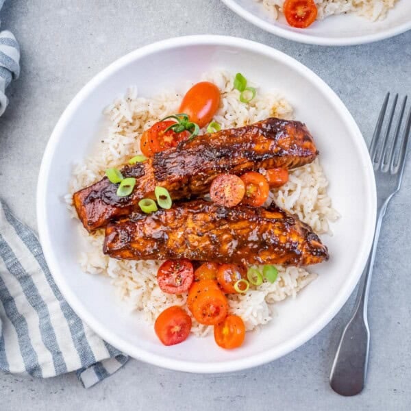 A white bowl with rice topped with 2 salmon fillets and tomato salad garnish.