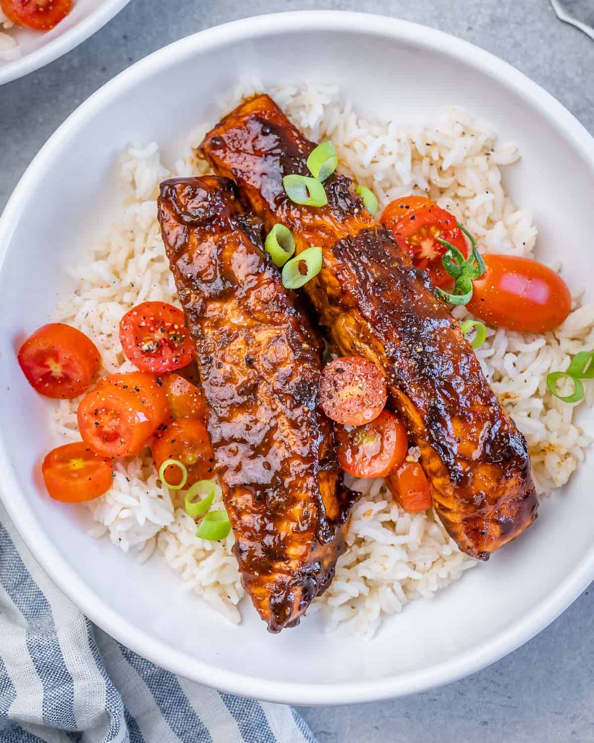 A white bowl with rice topped with 2 salmon fillets and tomato salad garnish.