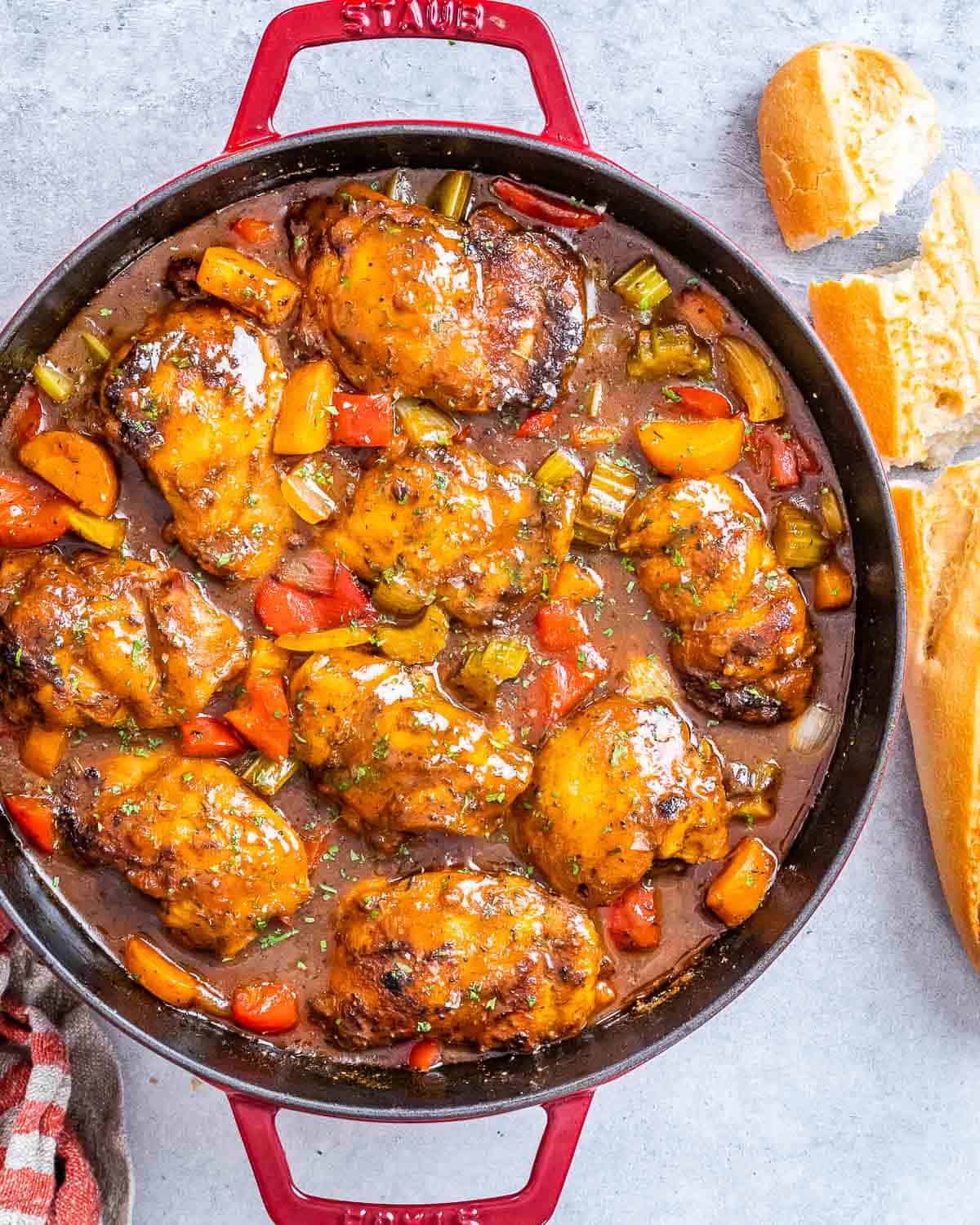 A red pot with brown stew chicken and a torn baguette on the side.
