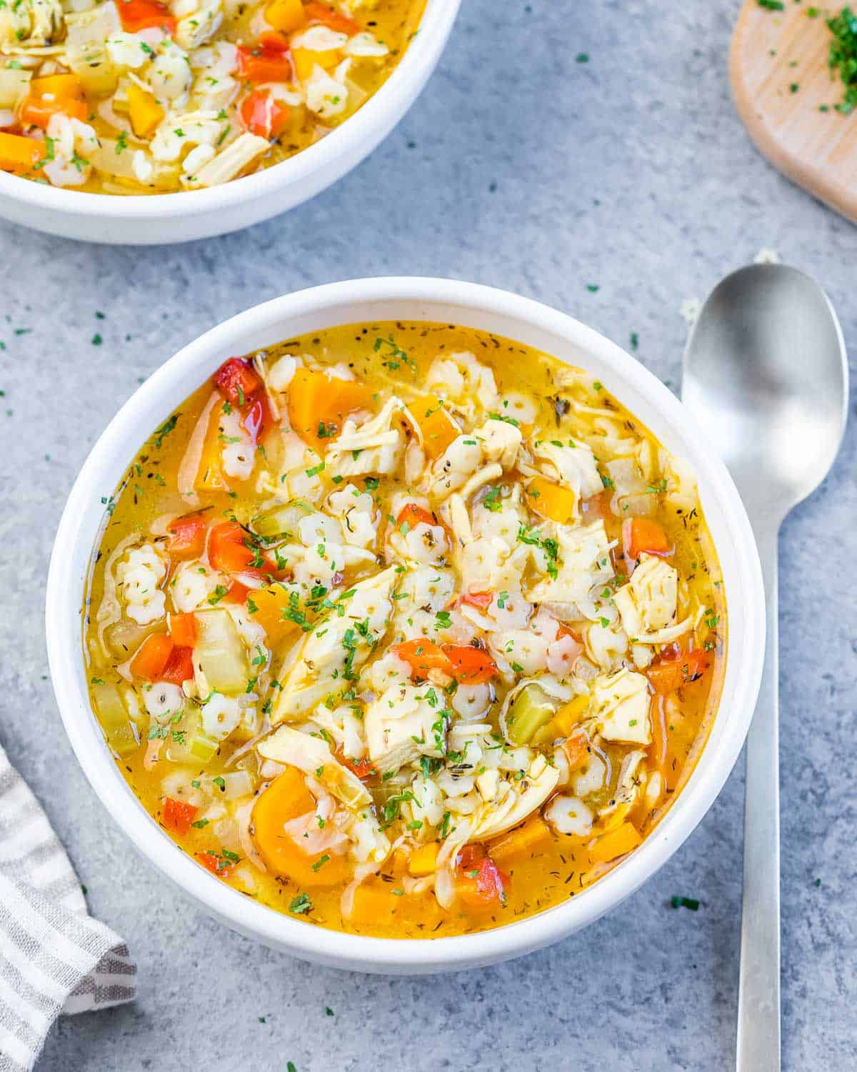 A white bowl of chicken pastina soup with a striped napkin and spoon.