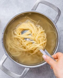 Hand lifting a portion of cooked spaghetti out of a pot of boiling water using kitchen tongs.