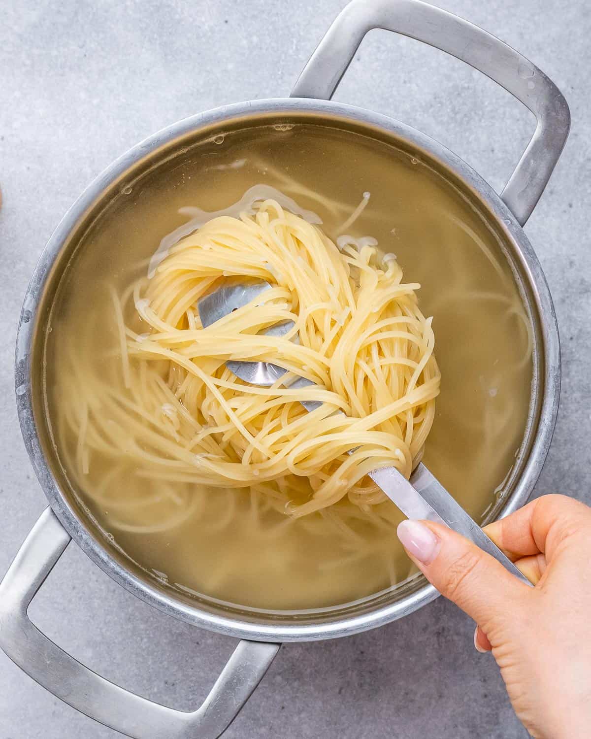 Hand lifting a portion of cooked spaghetti out of a pot of boiling water using kitchen tongs.